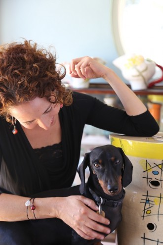 Kate Rosenberg resting her elbow on one of her hand-built stools, with her dachshund Oliver and her hand-built jugs in the background along with her collection of work by Imiso Ceramics and Light from Africa/Art in the Forest. Image: Inness Mass.